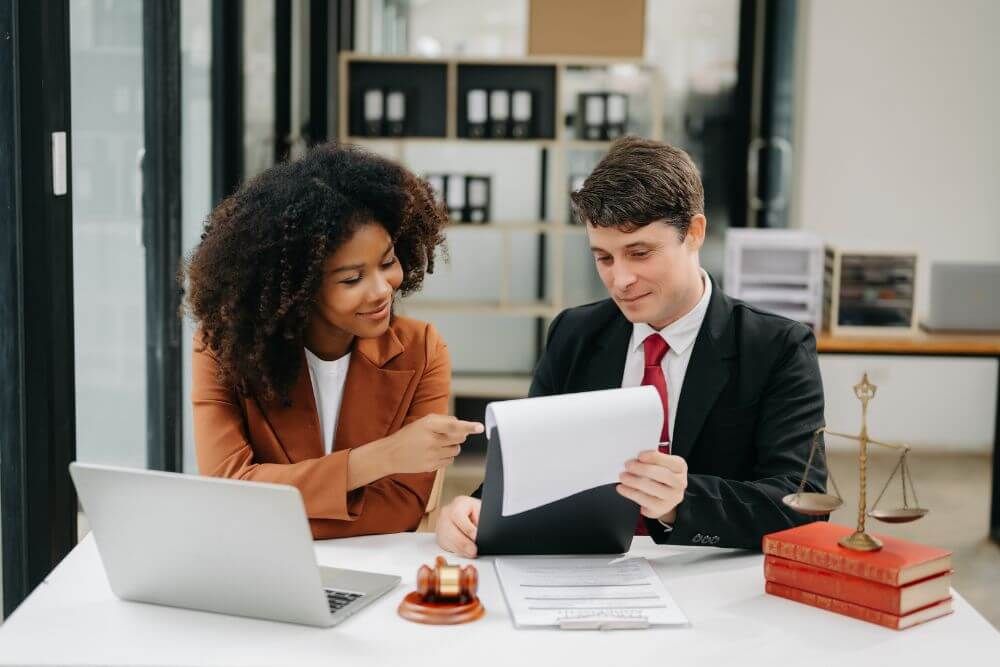 A woman with curly hair engages with a man in a suit, pointing at a document. They sit at a table with a laptop, books, and a gavel in a modern office.