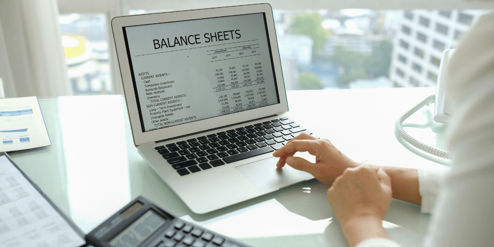 A person views a balance sheet on a laptop, with detailed financial data displayed. Nearby, a calculator and documents lie on a desk, set against an office cityscape backdrop.