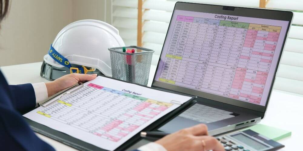 A person in professional attire reviews a costing report on a laptop while holding a clipboard. A safety helmet and a desk organizer with pens are visible in a well-lit office space.