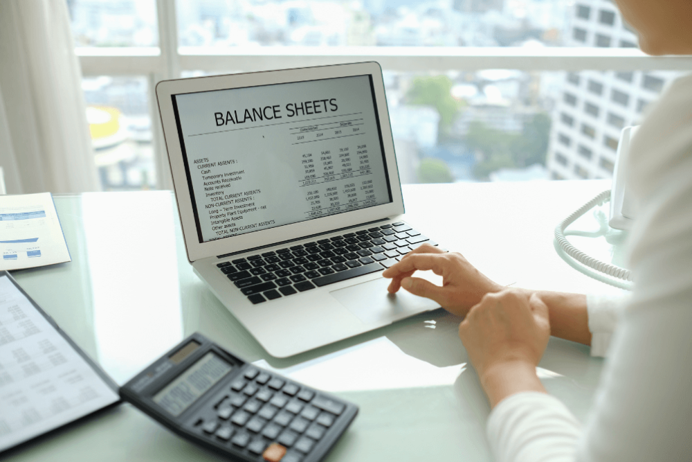 A person views a balance sheet on a laptop, with detailed financial data displayed. Nearby, a calculator and documents lie on a desk, set against an office cityscape backdrop.
