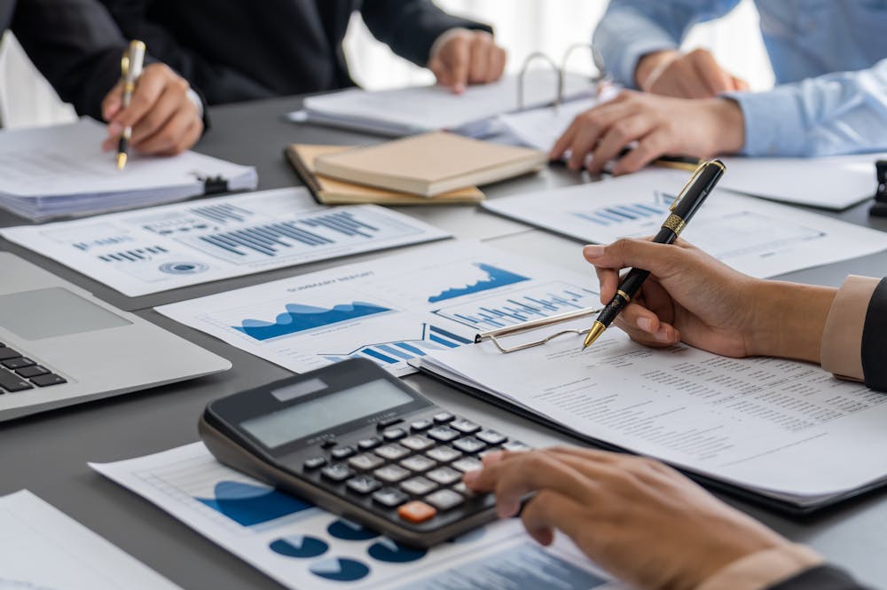 Hands holding pens write on financial documents and graphs spread across a table, surrounded by a laptop, calculator, notebooks, and other people in a business meeting context.