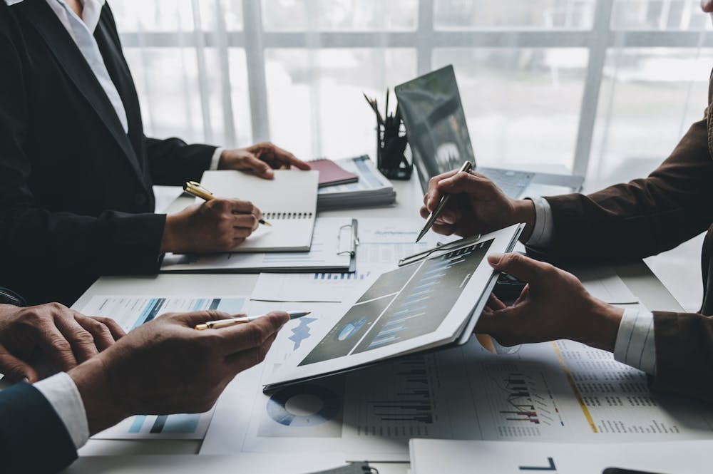 Business professionals discuss financial data displayed on a tablet, with papers, charts, and notebooks on the table in a modern office setting.