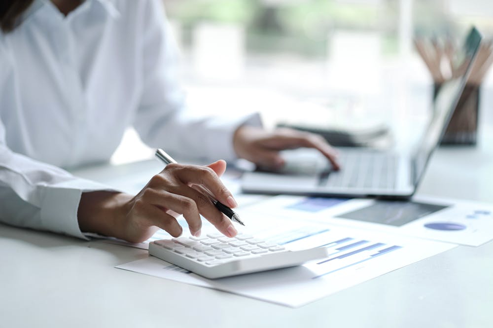 A person uses a calculator and pen while working on documents, with a laptop nearby in a bright office setting.