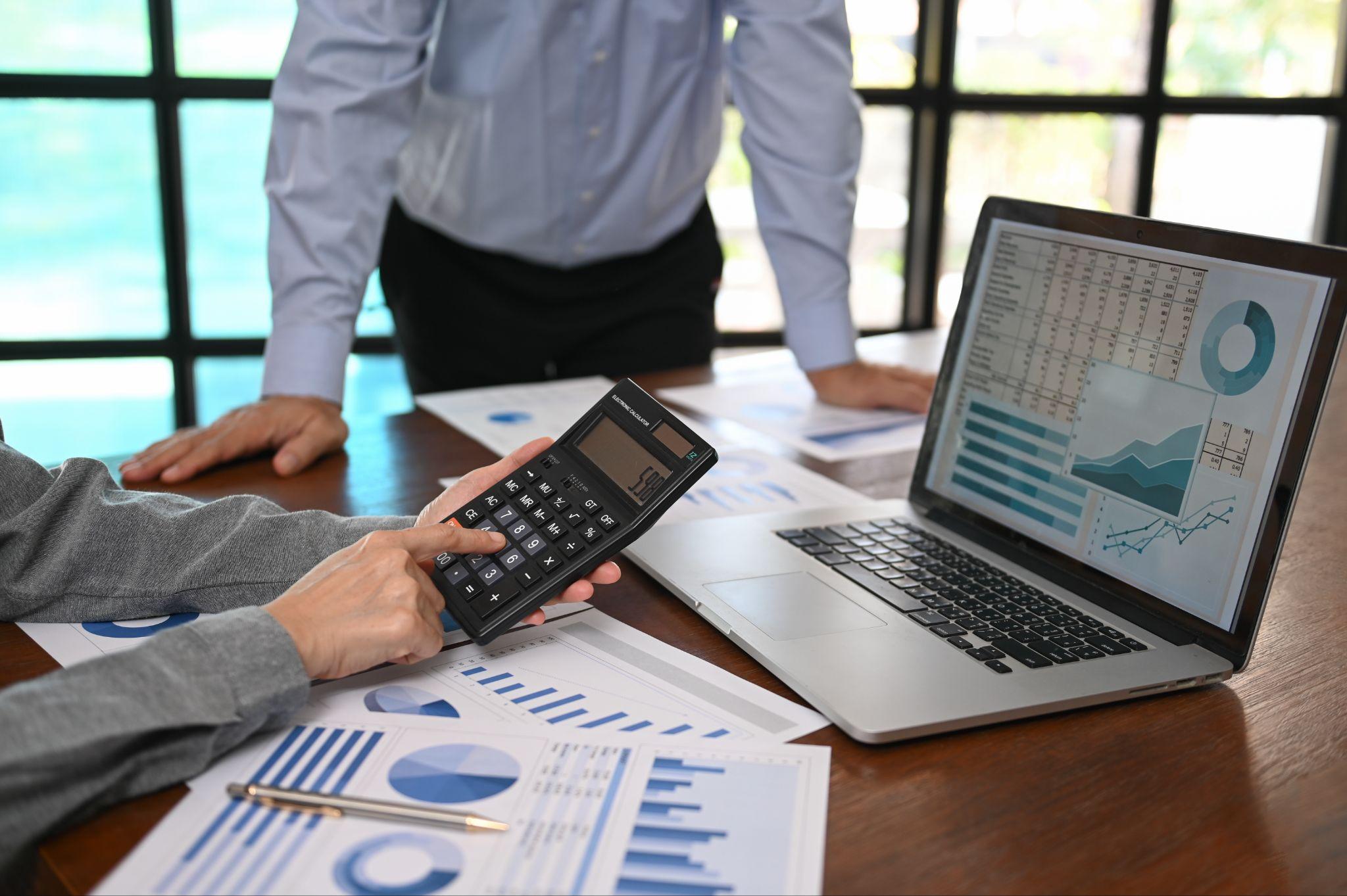 a person holding a calculator in front of a laptop, there another person standing, both are discussing metrics from different sources