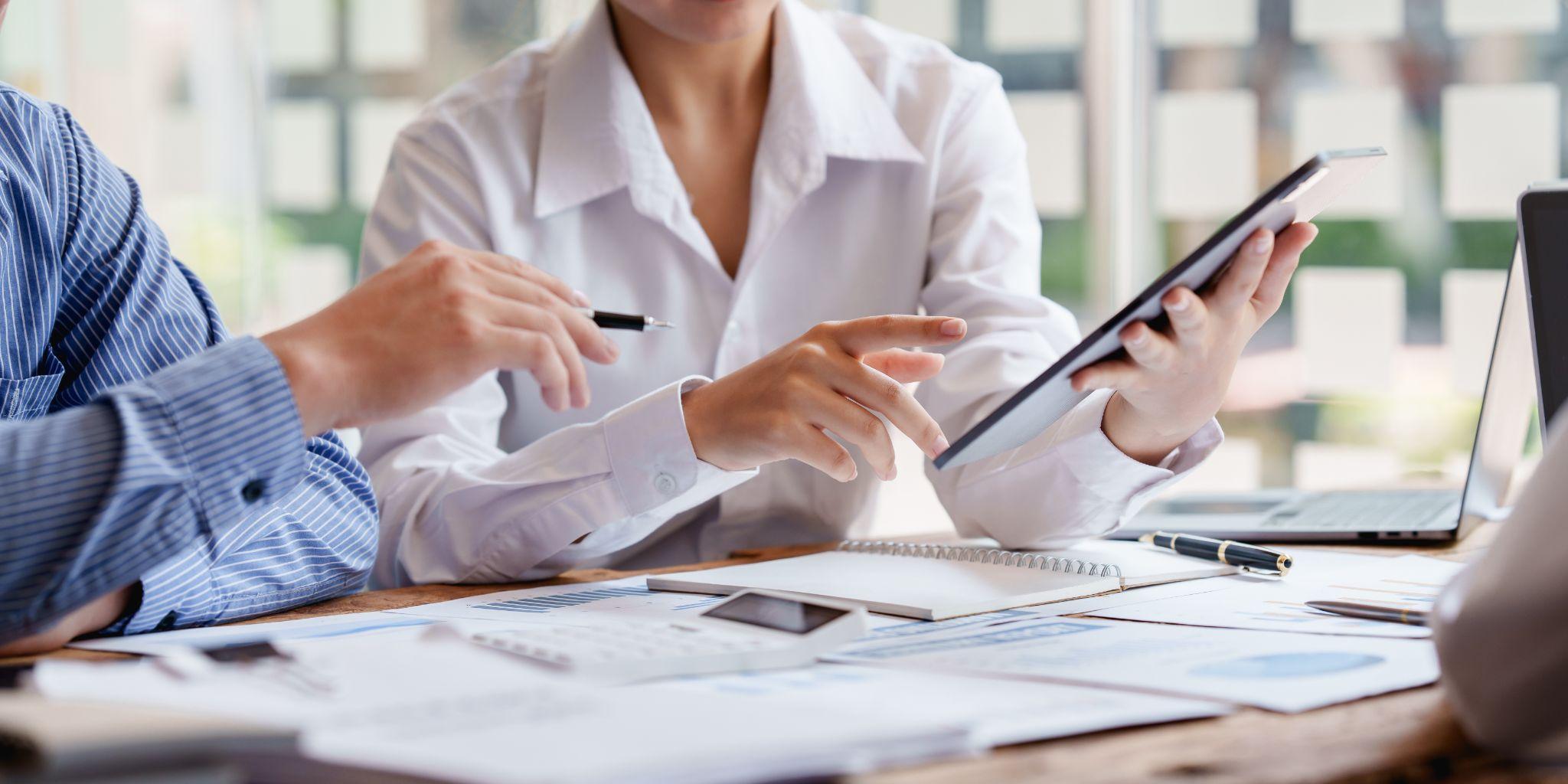 two people sitting at a table with a tablet, there are notebooks and papers