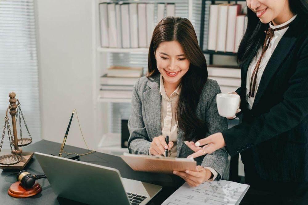 two women sitting at a desk in front of a laptop