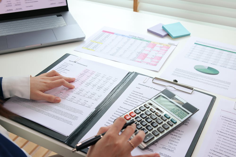 A hand uses a calculator to perform calculations on financial documents laid out on a table. A laptop and various charts and sticky notes are visible in the background.